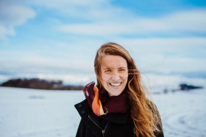 Portrait von Catalina Agudin. Sie befindet sich in einer Schneelandschaft, trägt eine dunkle Jacke und langes braun-blondes Haar. Sie lacht. Der Himmel ist blau und mit vielen weissen Wolken bedeckt.